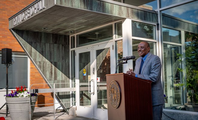 A man in a suit stands at a podium in front of the Irvin D. Reid Hall entrance.