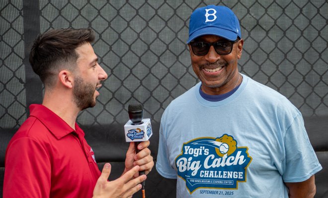 Man in red shirt interviews a man in sunglasses and a blue hat.