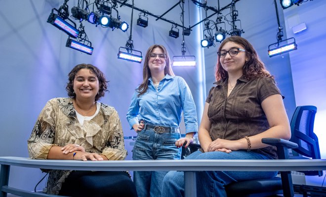 In a newsroom, three women, two seated at a desk and one standing between them pose with studio lights shining behind them.