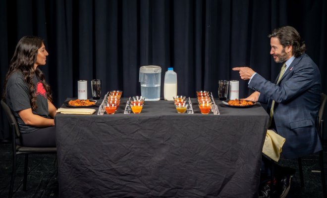 A man in a suit and tie gestures to a woman, both of whom are sitting in front of plates of chicken wings.