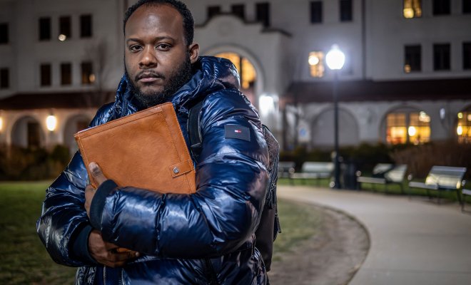 Horatio Wauchope , wearing a winter coat, stands outside on a campus walkway at night, holding a large folder to his chest in front of a lit academic building.