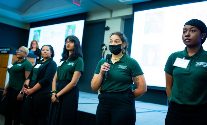 Five students in green polo shirts in front of projectors giving presentation
