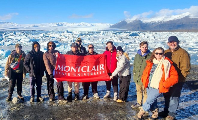 A group of Montclair students and professors stand together on a glacier in Iceland, holding a red Montclair State University banner. They are dressed in cold-weather gear, surrounded by a rugged, icy landscape. In the background, the glacier stretches into the distance, with patches of snow and exposed rock.