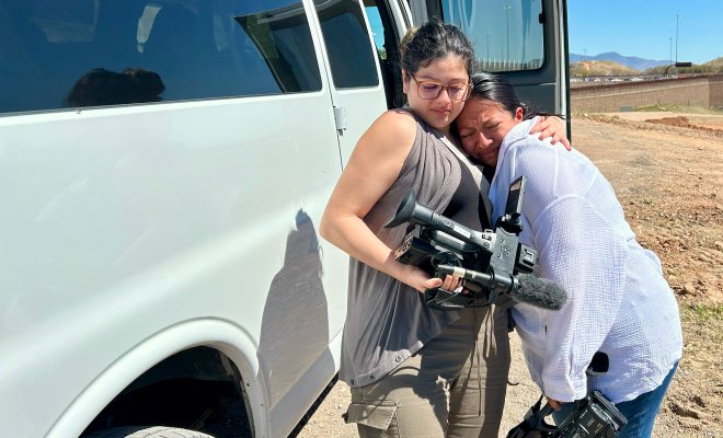 A student holding a video camera hugs another student in the desert.