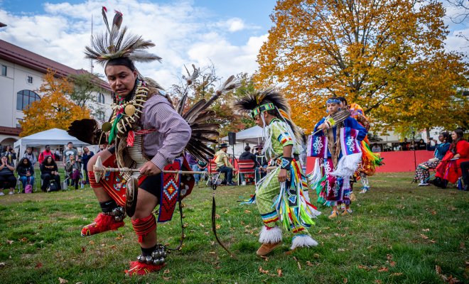 Native American men and women in traditional costumes dance outdoors while spectators watch.