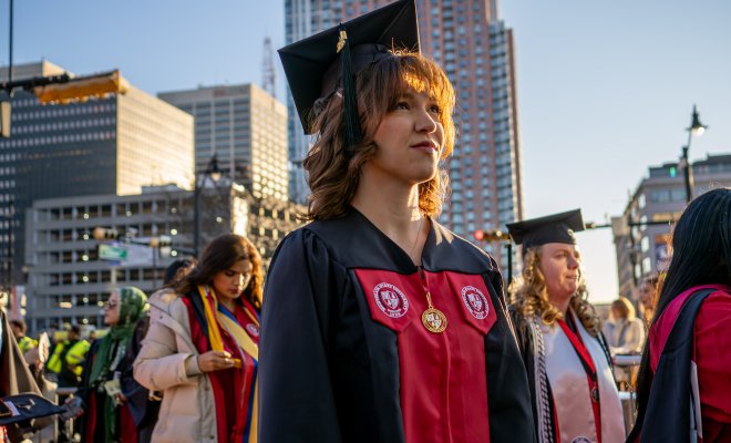 A student standing outdoors in the sun wearing a robe and mortar board at commencement looking up 