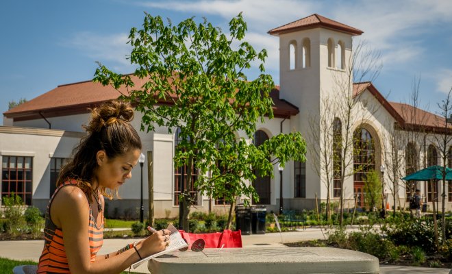 A student sitting at a table outside and writing something in a notebook. Montclair's School of Communication and Media in the background.