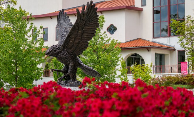 This image shows a statue of a red hawk with some red flowers in the foreground.