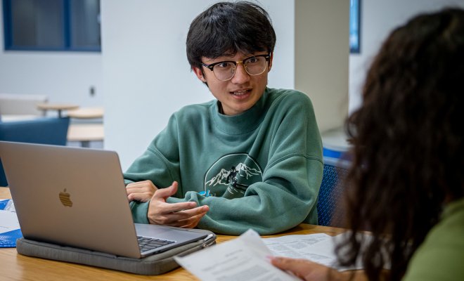 Student wearing green sweatshirt and glasses talks to another student across a table with his laptop open.