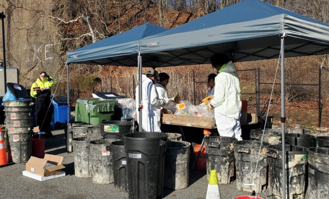 Photo of the staff from SCS Engineers as they stand around a sorting table performing a waste audit. They are dressed in personal protective equipment and surrounded by empty waste bins to collect the various types of waste they sort out.