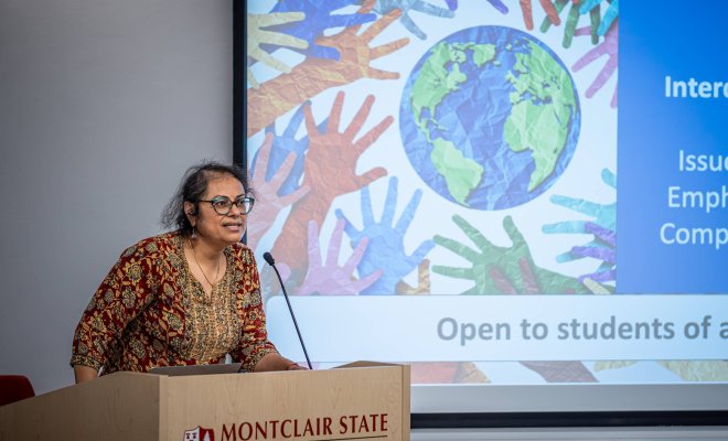 A college professor stands in front of a PowerPoint and addresses an audience