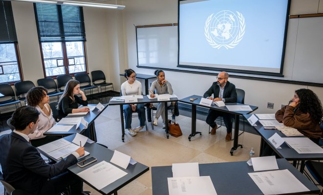 College students sit in a circle around a professor, the United Nations logo appears on the screen behind them