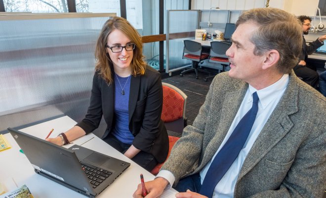 Two people sit around a laptop at a desk in an office environment