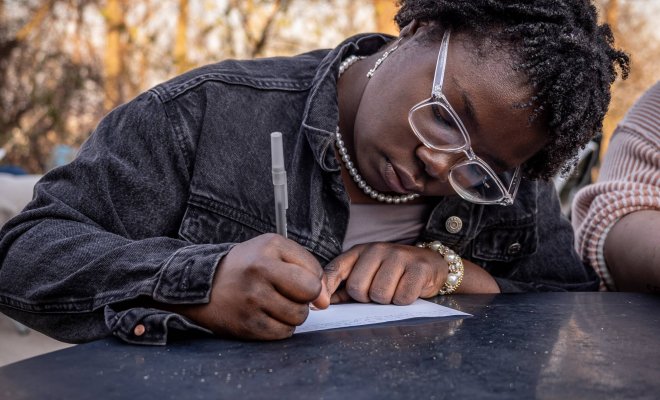 A student leaning over a table, writing with a pen on a piece of paper