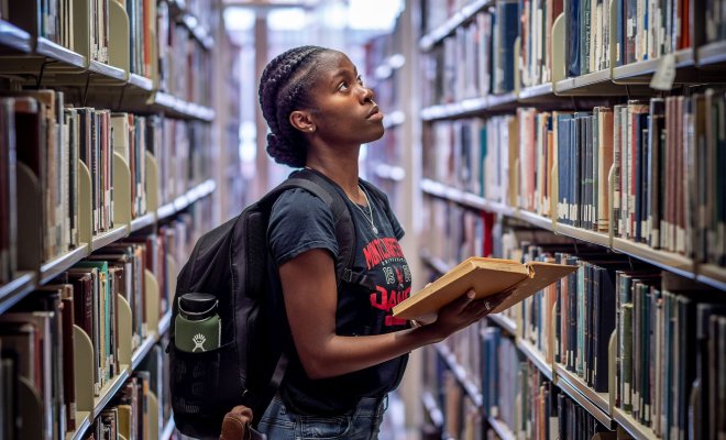 A student stands in the middle of a library aisle, holding a book and studying the shelf