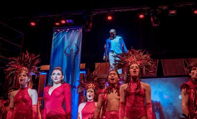 Performers in red costumes singing on stage during a production of Pippin from 2022