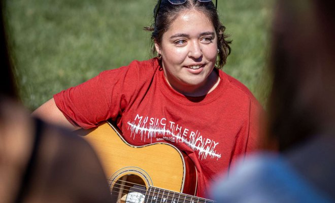 A student holds a guitar while seated in the grass.