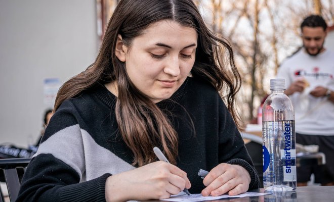 A college student sits at an outdoor table and writes on a piece of paper