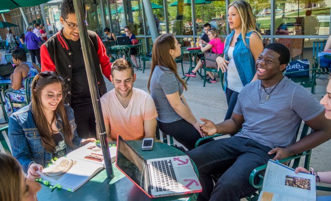 A group of college students sit around an outdoor table under an umbrella with laptops and books out