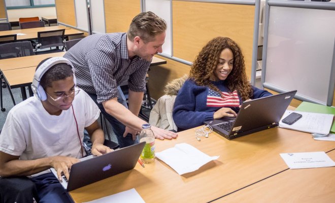 Two college students sit in front of laptops at a desk as an instructor leans over their shoulder 