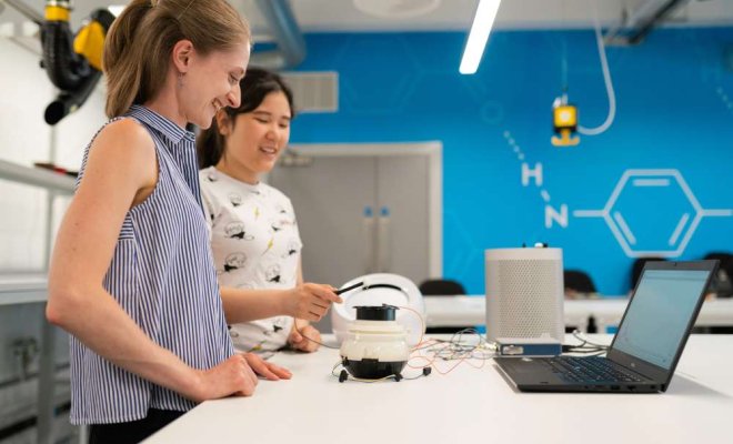 two women stand in a STEM engineering lab with computer on table and bot infront of them