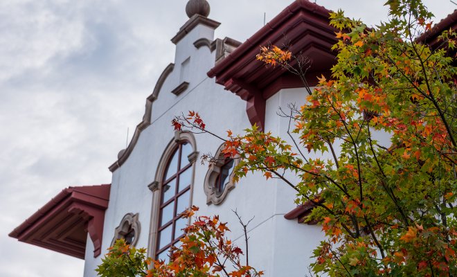 A campus building with Spanish-style architecture is framed by colorful autumn trees under a cloudy sky.