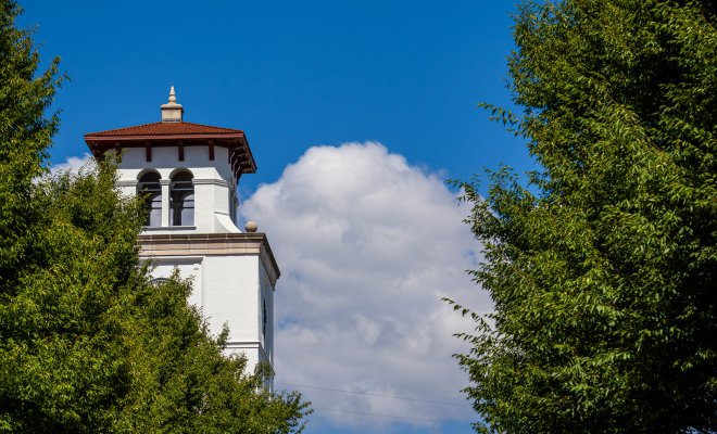 A white campus bell tower rises above green trees beneath a vivid blue sky with large white clouds.