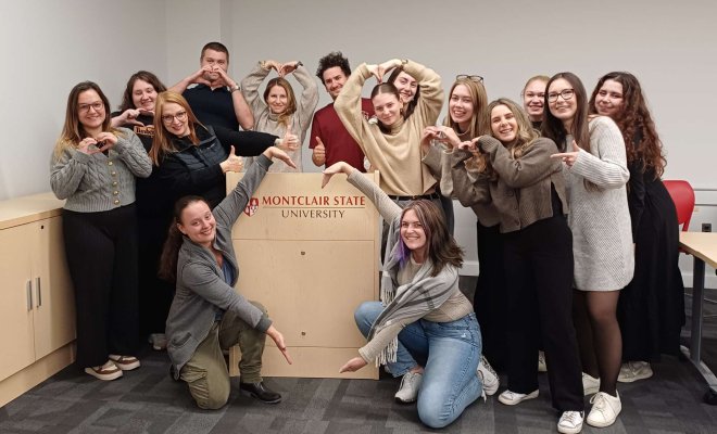 A group of people pose around a podium and point to the "Montclair State University" logo on it