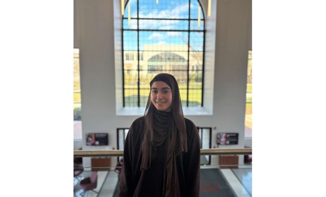 A woman wearing a hijab poses for a headshot in front of a window 