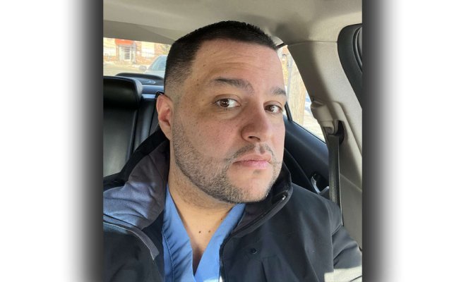 Headshot of a man wearing a blue shirt and a black jacket sitting inside a car