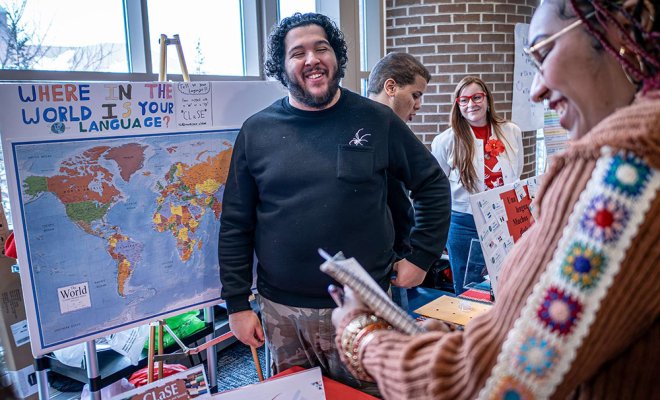 A man stands in front of a world map at a booth and smiles at a visitor