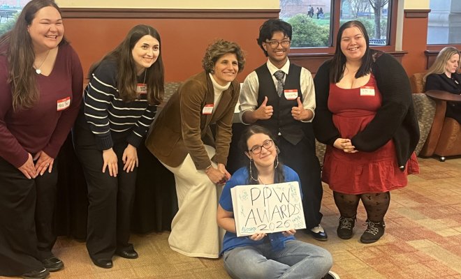 A group of six college students pose in a line, one students sits and holds a book which has the words "PPW Awards 2026" written in it 