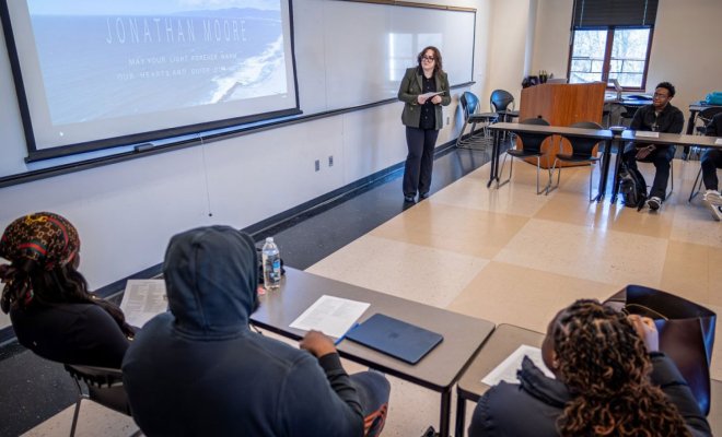 A college student stands in front of a projector screen and presents to a class full of students