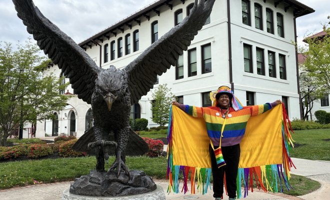College student wearing a rainbow-colored fringed robe, rainbow sweater, colorful hat, and rainbow shoes, standing in front of a bronze statue of a Red Hawk.
