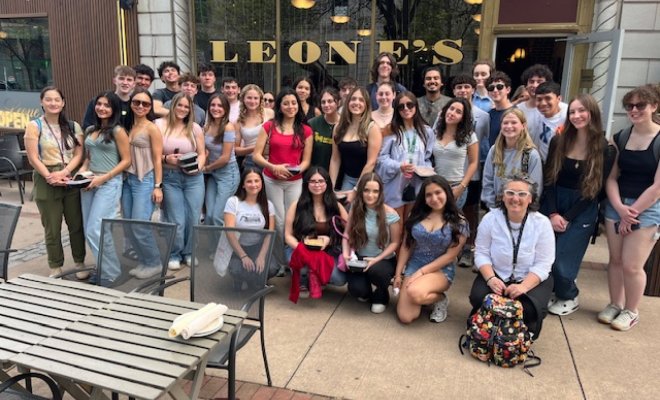 A large group of college students pose on a sidewalk outside an Italian restaurant