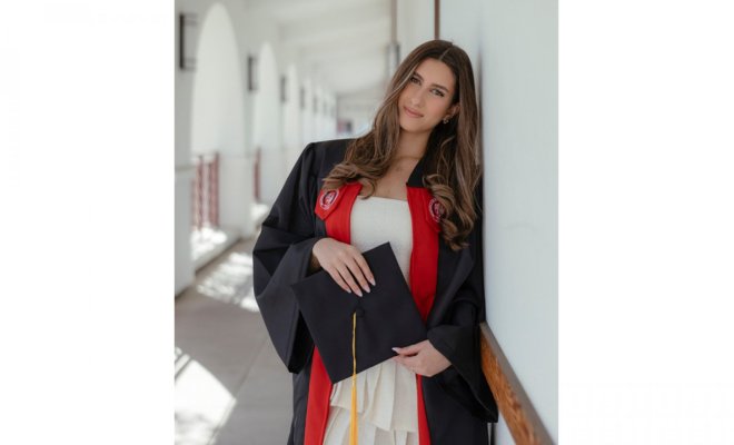 A college student in a graduation robe holds a mortarboard and leans against the wall of a courtyard