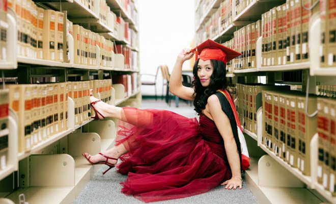 A college student dressed in a red cap and gown sits on the floor between shelves in a library