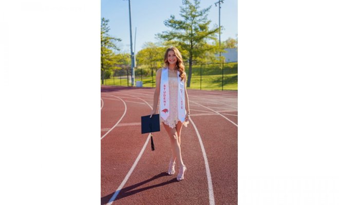 A college student stands holding a mortarboard on an outdoor running track