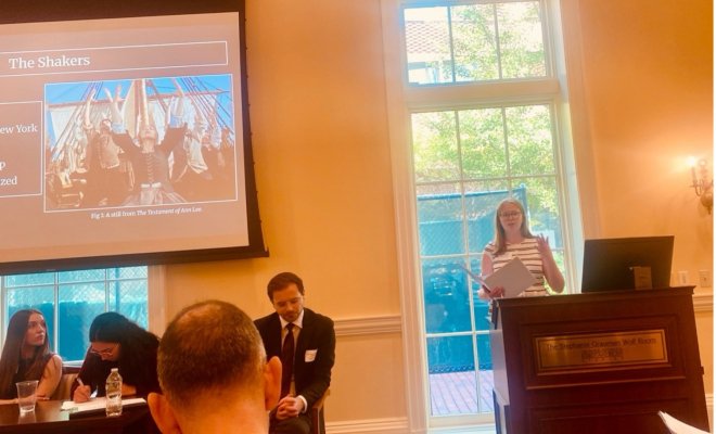 A woman stands at a dais and presents a PowerPoint to a room full of people