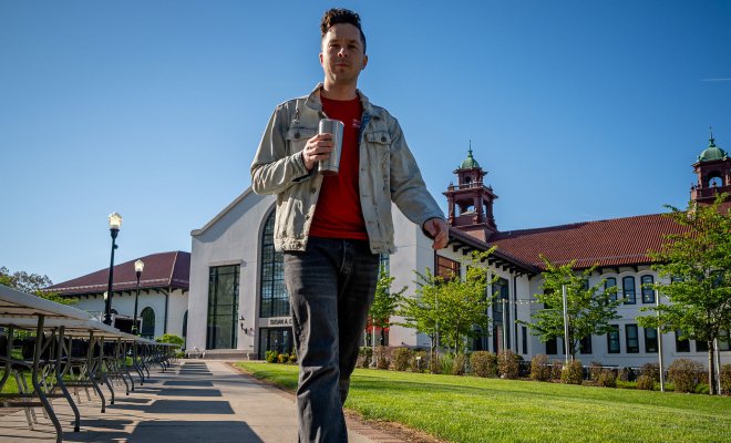 Casey Coleman walks along a campus pathway, holding a travel coffee mug, with long rows of empty folding tables on one side.