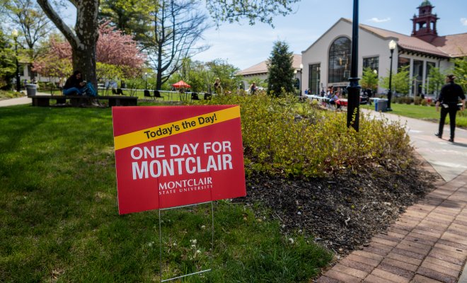 A red sign with while lettering says: One Day for Montclair. The campus is in background.