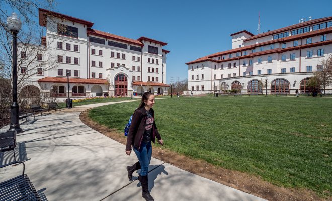 A student walking on campus in front of some buildings.