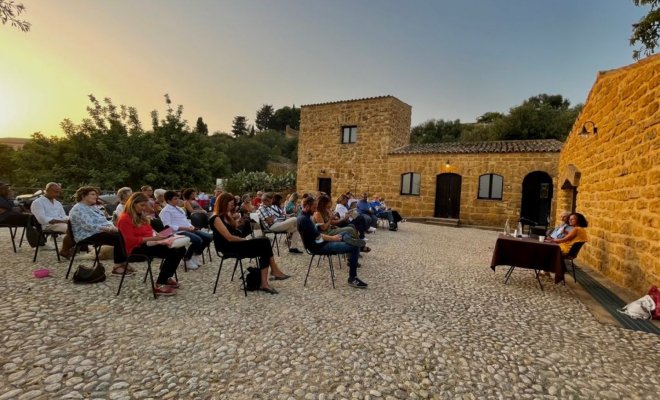 A woman sits at an outdoor table in a stone courtyard and speaks to an audience