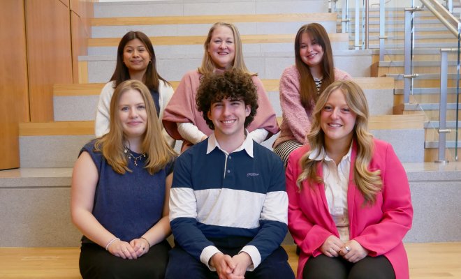 Six people dressed professionally in two rows of three sitting on stairs. They are smiling.