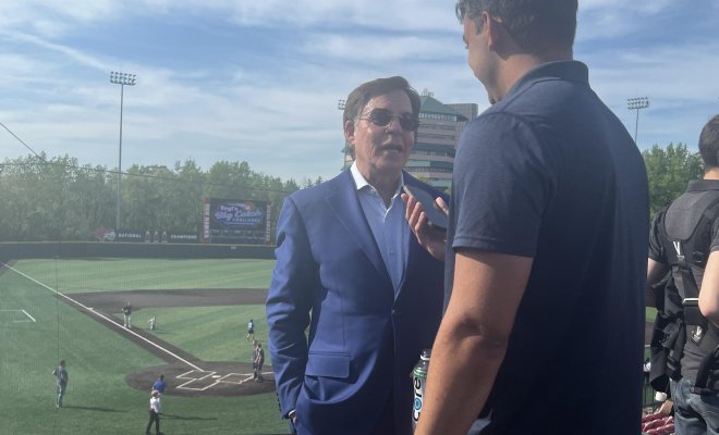 wo people standing outside in an empty sports stadium. The on in the foreground is interviewing the other person using is mobile phone.