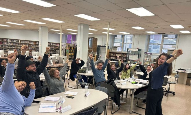 adult students seated at four tables in a library, stretching arms upward following the teacher at the front of the room