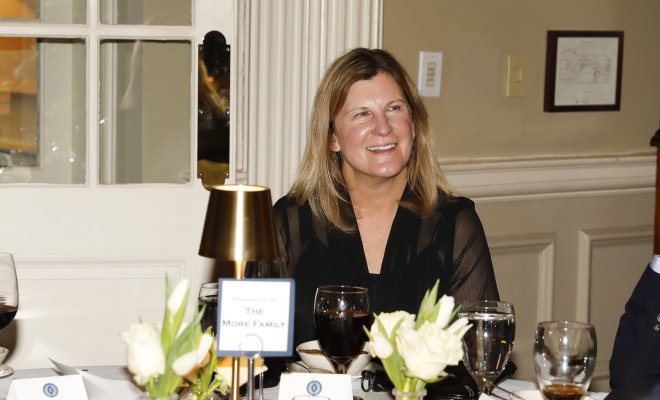 A woman with long dirty blonde hair sitting at a table. She is smiling and happy.