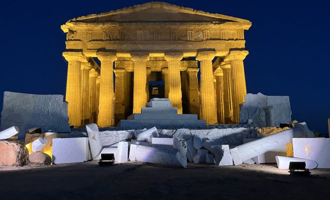A performance stage at night with fake crumbled marble ruins and an image of the Parthenon behind it 