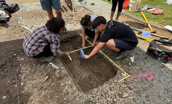 Three college students bend over an excavation and hold a measuring tape