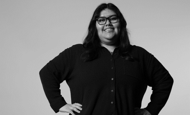 A black and white photo of a woman with long hair and glasses. She is smiling, standing and with her hands on her hips.